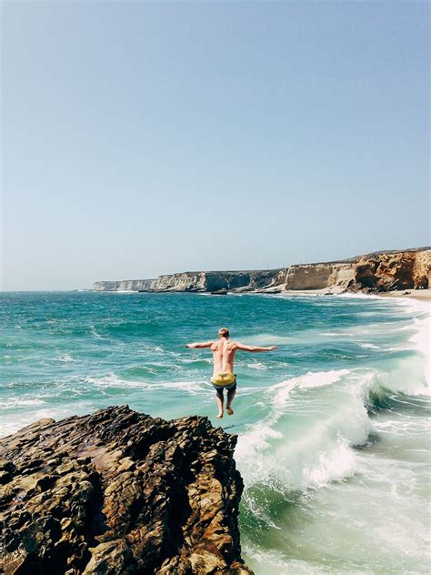 Man Jumping From A Rock Cliff Into The Ocean Waves With His Arms Stretched Out By Stocksy