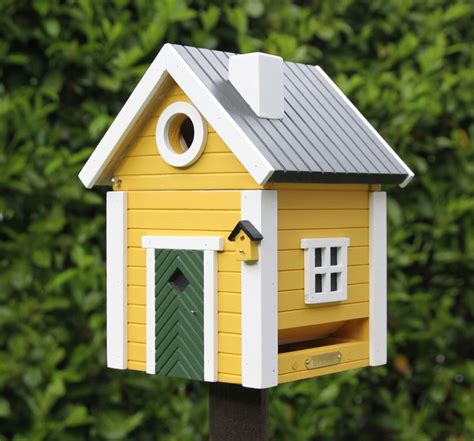 A Yellow Bird House Sitting On Top Of A Wooden Post In Front Of Some Bushes