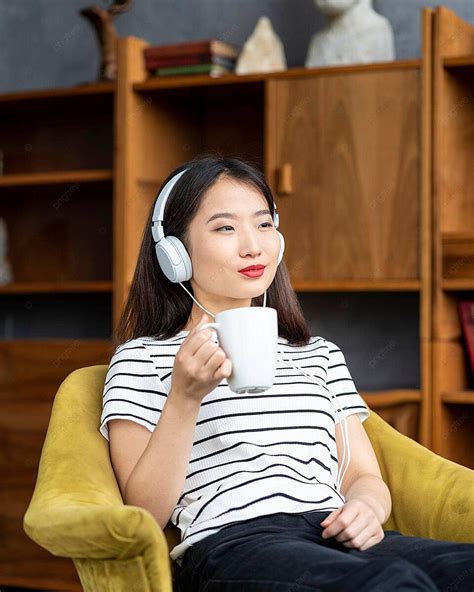 Charming Asian Lady Enjoying Music With Headphones While Seated On A Chair Photo Background And