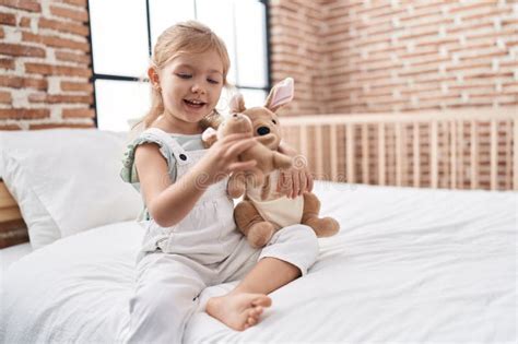 Adorable Blonde Girl Playing With Doll Sitting On Bed At Bedroom Stock