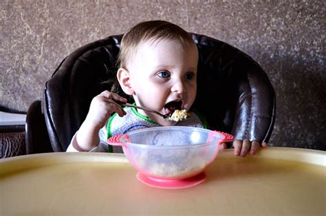 Baby Girl Eating Porridge While Sitting In Baby Chair Stock Photo