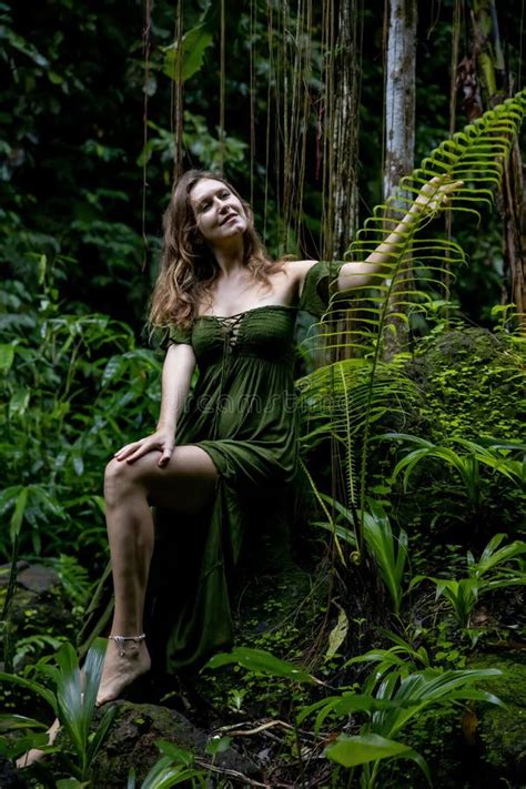 Woman In Jungle Sitting On A Stone Surrounded By Lush Green Plants Touching Fern Leaf