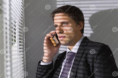 Businessman Peeking Through Blinds While On Call In Office Stock Image