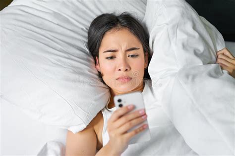 Close Up Portrait Of Asian Girl Lying In Bed Looking At Smartphone Concerned Waking Up Late