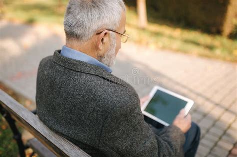 Grandpa Uses A Tablet Sitting In The Park On The Bench Stock Photo
