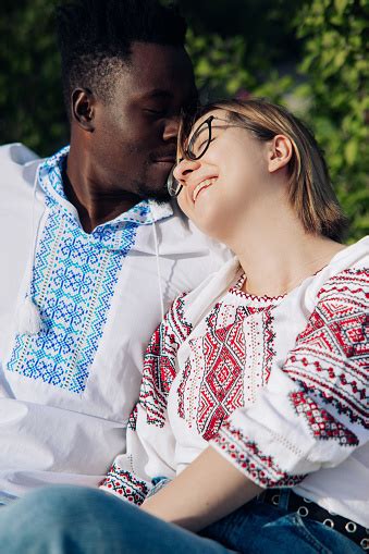 Interracial Happy Couple Sits On Bench In Garden Dressed In Ukrainian Embroidered Shirts Stock