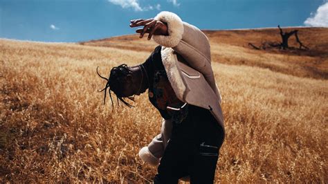 Travis Scott Is Looking Down Standing On Dry Grass In Sky Background