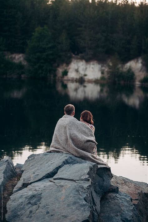 Two People Sitting On Top Of A Rock Next To A Body Of Water With Trees In The Background