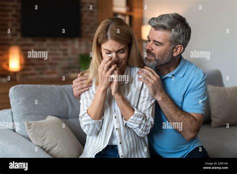 Sad Middle Aged Caucasian Man With Beard Hug Calm Crying Unhappy Wife In Living Room Interior