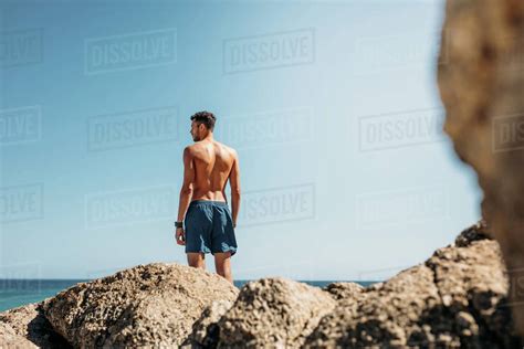 Rear View Of A Shirtless Man Standing On Top Of Rocks Near The Sea