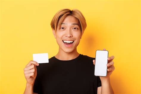 free photo amazed happy asian guy showing credit card and smartphone screen smiling