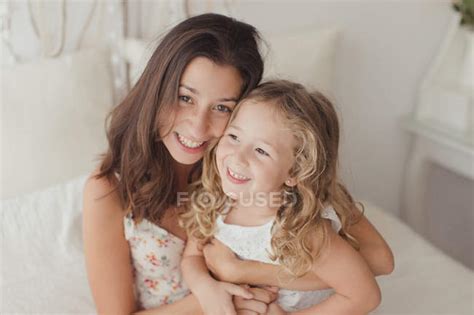 happy brunette mother having fun with blonde cute daughter while embracing on bed — looking in