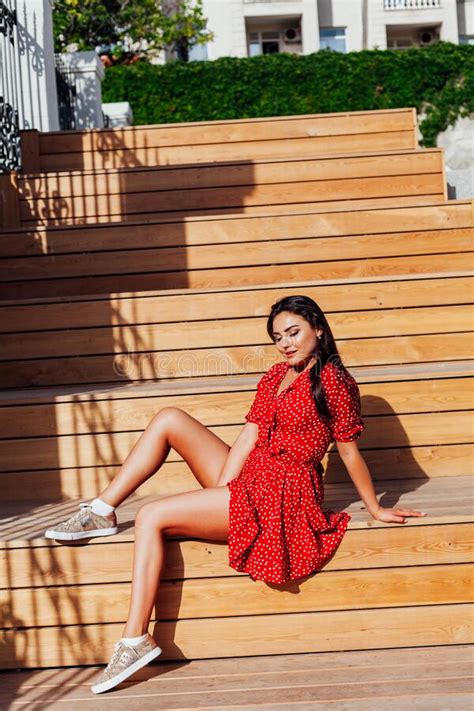 Beautiful Brunette Woman In Red Dress In Polka Dots Sits On A Bench