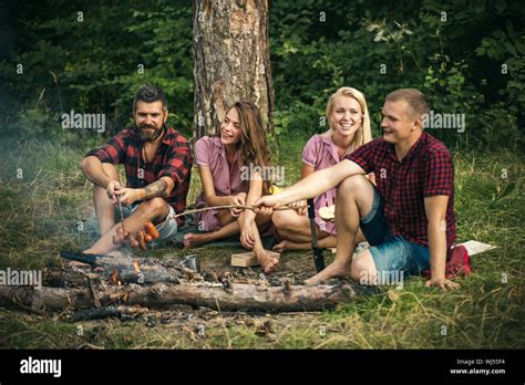 two couples camping in woods guys frying sausages over fire smiling friends sitting around