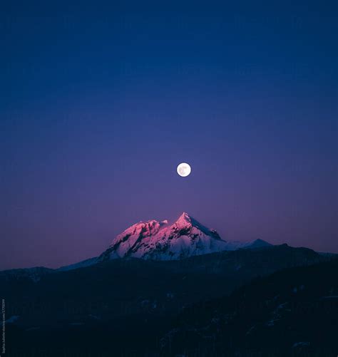 Moon Rising Behind The Peak Of A Snow Covered Mountain By Stocksy