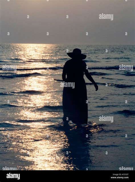 Silhouette Of Woman In Dress And Hat Wading Through The Shallow Water