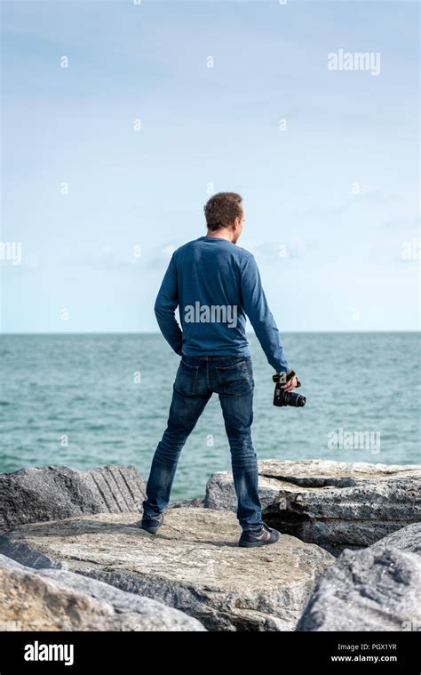 Man Standing On Rocks Looking Out To Sea Holding A Camera Back View