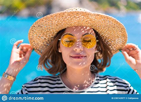 Pretty Woman In Yellow Sunglasses And Straw Hat Smiling Sincerely To Camera On Teal Blue Sea