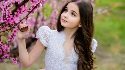 Little Cute Girl Is Standing Near Pink Blossom Tree Wearing White Dress