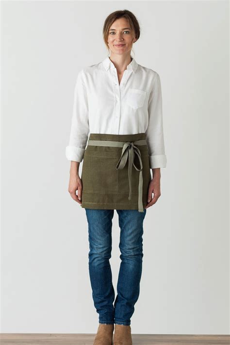 a woman standing in front of a white wall wearing an apron and jeans with her hands on her hips