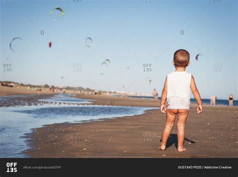 Back View Of Baby With Naked Bottom Standing On Beach Watching Flying Parachutes Stock Photo