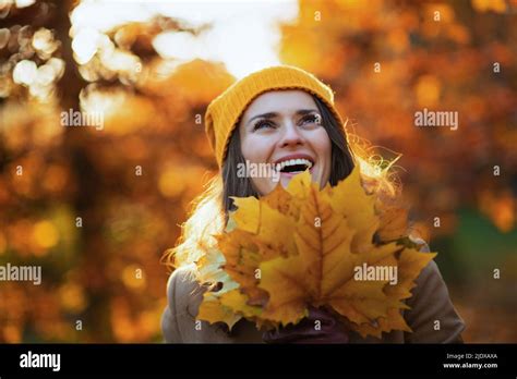 hello autumn smiling middle aged woman in beige coat and orange hat with autumn yellow leaves