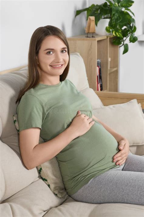 Happy Pregnant Woman Sitting On Sofa And Touching Her Belly In Living