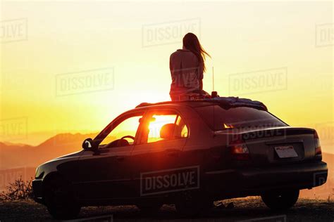 Rear View Of Woman Looking At View While Sitting On Car Roof Against