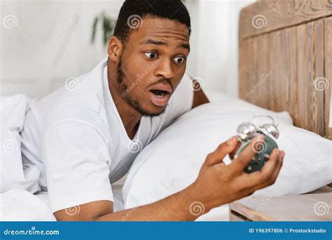 Overslept African Man Holding Alarm Clock Waking Up In Bed Indoor Stock