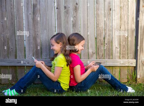 Twin Sister Girls Playing With Tablet Pc Sitting On Backyard Lawn Fence