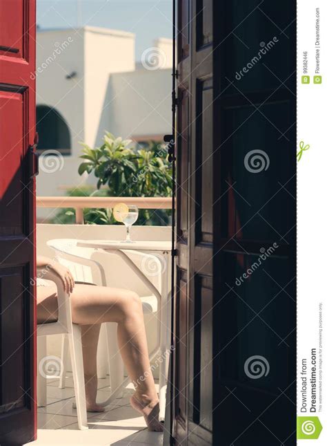 Female In A Swimsuit Sits On The Balcony Of An Old Greek Building With Lemon Water In A Glass