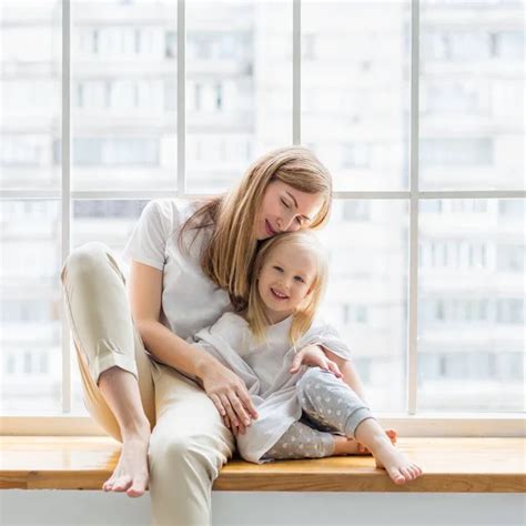 Happy Mother Hugs Her Daughter While Sitting In Front Of Window Stock
