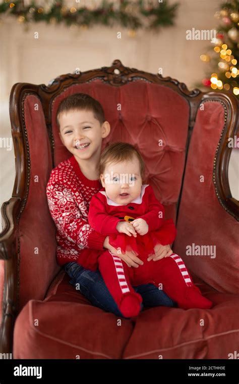 litttle brother hugs his little sister in red retro chair near christmas tree enjoying a love