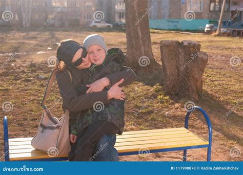 Mom Kisses And Hugs Her Son Sitting On A Bench In The Courtyard Of A