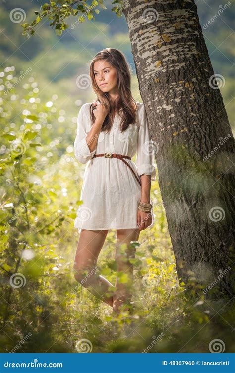 attraktiv ung kvinna i den vita korta klänningen som poserar nära ett träd i en solig sommardag