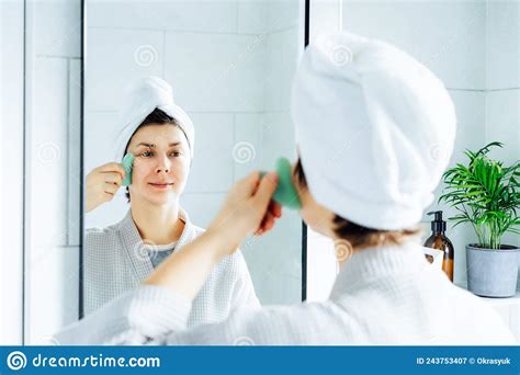 Woman In A Bathrobe With A Towel On Her Head Making Face Massage With Gua Sha Roller In The