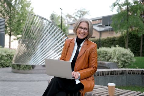 pretty gray haired business woman of mature age works with a laptop on a bench in the park stock