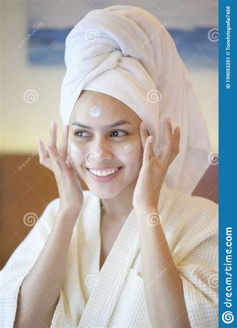 Happy Beautiful Woman In White Bathrobe Applying Moisturizing Cream On Face In Bedroom Skin