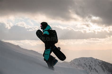 Side View Of Man In Ski Suit With Snowboard Walks At Snow Covered Slope