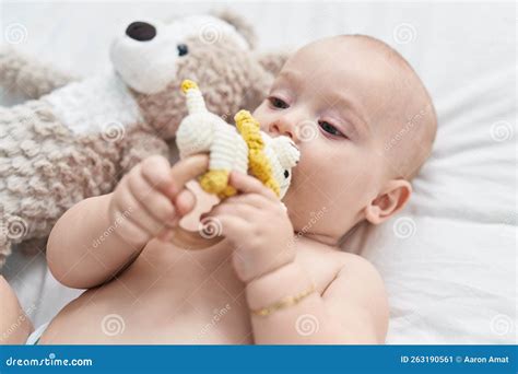 Adorable Caucasian Baby Lying On Bed Biting Doll At Bedroom Stock Image
