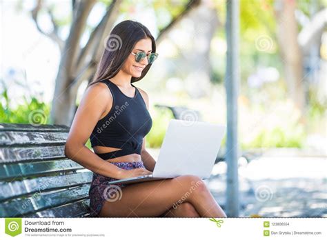 Pleased Brunette Latin Woman Isitting On Bench In Park And Using Laptop