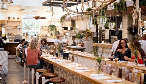 People Sitting At Tables In A Restaurant With Plants Hanging From The Ceiling And Potted Plants