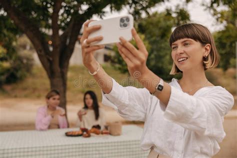 Attractive Young Caucasian Woman Takes Selfie Against Backdrop Of Sitting Friends In Nature Day