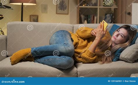 Tired Woman Texting On Smartphone While Lying On A Sofa In The Living Room Smiling Blonde Girl