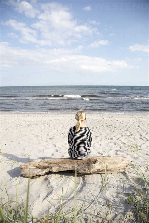 Mature Woman Sitting On Driftwood And Looking At Sea Stock Photo