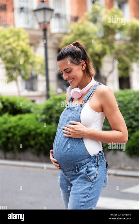 Side View Of Dreamy Pregnant Female In Overalls Standing On Street While Enjoying Stroll And