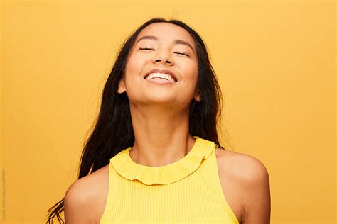 happy asian woman portrait with eyes closed laughing isolated over yellow by stocksy