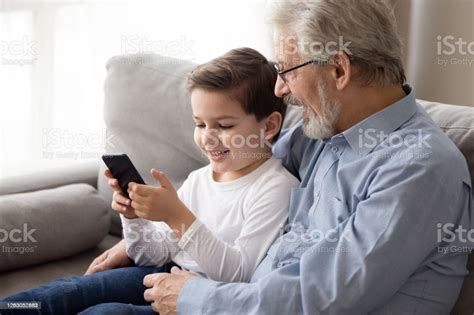 Elderly Grandfather Embraces Grandson Holding Smartphone Using Gadget