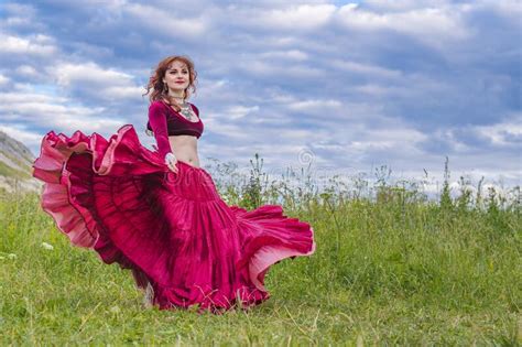 Young Red Haired Girl In Red Dance Dress Dances On Green Meadow Against