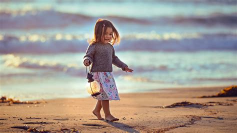 Cute Little Girl With Lantern Is Walking On Beach Sand Wearing White